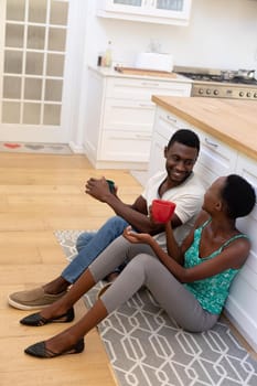 African american couple sitting on floor in kitchen holding mugs. staying at home in isolation during quarantine lockdown.