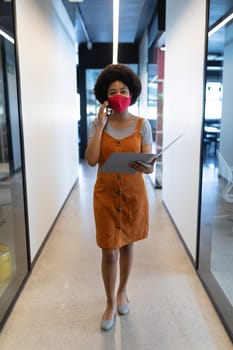 Mixed race businesswoman wearing face mask in creative office. woman talking on smartphone and holding documents. social distancing protection hygiene in workplace during covid 19 pandemic.
