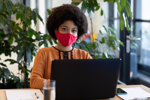 Mixed race businesswoman wearing face mask in creative office. portrait of a woman looking at camera and using laptop. social distancing protection hygiene in workplace during covid 19 pandemic.