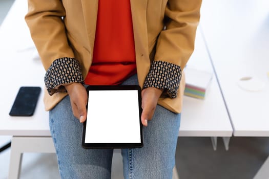 Midsection of mixed race businesswoman sitting on desk using tablet in creative office. social distancing in workplace during covid 19 pandemic.