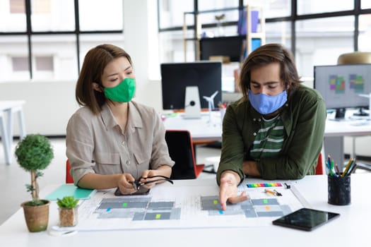 Diverse male and female colleague wearing face masks looking at blueprints and discussing. health protection hygiene in creative office during covid 19 pandemic.