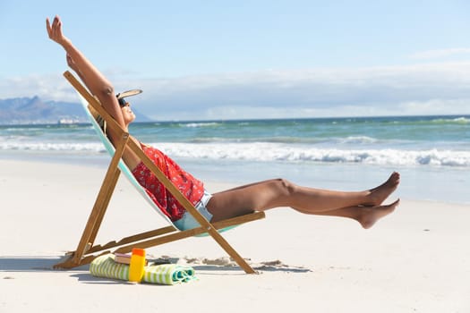 Mixed race happy woman on beach holiday sitting in deckchair stretching. healthy outdoor leisure time by the sea.