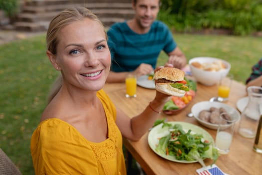 Portrait of smiling caucasian woman holding hamburger eating meal with family in garden. family celebrating independence day eating outdoors together.