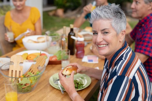 Portrait of smiling caucasian senior woman holding hotdog at table with family having meal in garden. three generation family celebrating independence day eating outdoors together.