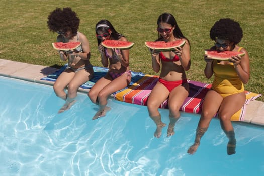 Diverse group of female friends eating watermelon sitting at the poolside. Hanging out and relaxing outdoors in summer.