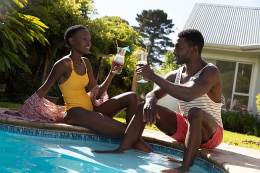 African american couple sitting at the poolside and drinking drinks on sunny garden terrace. staying at home in isolation during quarantine lockdown.