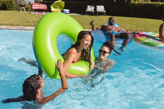 Diverse group of friends having fun playing on inflatables in swimming pool. Hanging out and relaxing outdoors in summer.