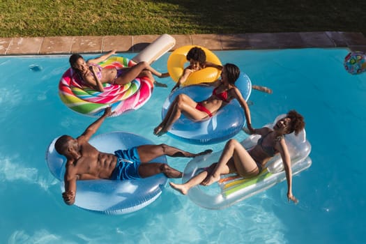 Diverse group of friends having fun playing on inflatables in swimming pool. Hanging out and relaxing outdoors in summer.