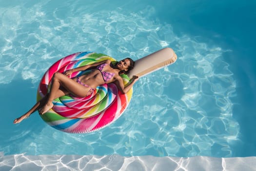 Mixed race woman sunbathing on inflatable in swimming pool. Hanging out and relaxing outdoors in summer.