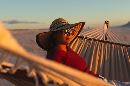 Mixed race woman on beach holiday sitting in hammock during sunset. healthy outdoor leisure time by the sea.