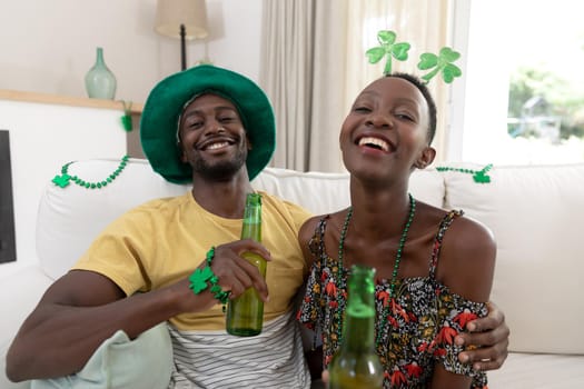 African american couple wearing st patrick's day costumes laughing and holding glasses of beer. staying at home in isolation during quarantine lockdown.