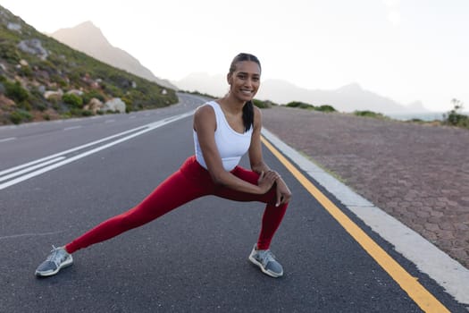 Fit african american woman in sportswear stretching on a coastal road. looking at the camera and smiling. healthy lifestyle, exercising in nature.