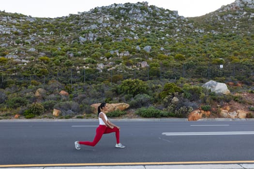 Fit african american woman in sportswear stretching on a coastal road. looking at the camera and smiling. healthy lifestyle, exercising in nature.