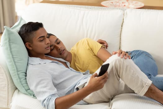 Diverse gay male couple sitting on sofa using smartphone and embracing. staying at home in isolation during quarantine lockdown.