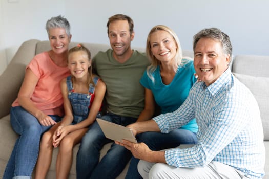 Smiling caucasian grandparents on couch with granddaughter and her parents looking at tablet. happy three generation family spending time together at home.