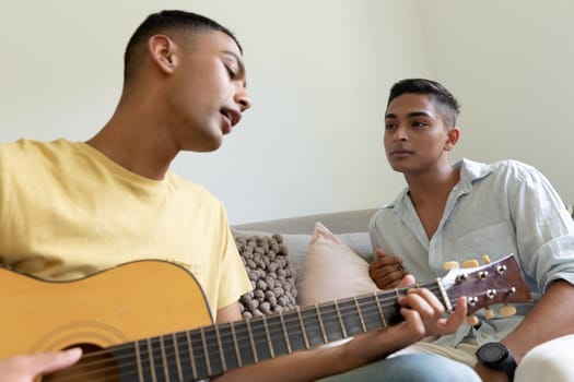 Diverse gay male couple sitting on sofa playing guitar. staying at home in isolation during quarantine lockdown.