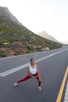 Fit african american woman in sportswear stretching on a coastal road. looking at the camera and smiling. healthy lifestyle, exercising in nature.