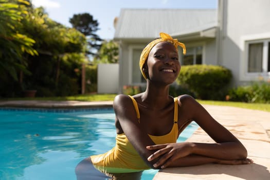 Portrait of african american woman in water leaning on poolside on sunny garden terrace. staying at home in isolation during quarantine lockdown.