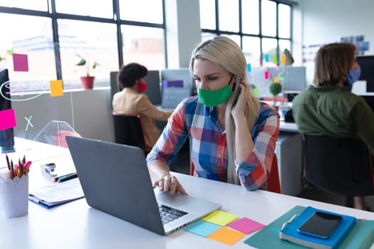 Caucasian businesswoman wearing face mask in creative office. woman sitting at desk, using laptop computer. social distancing protection hygiene in workplace during covid 19 pandemic.