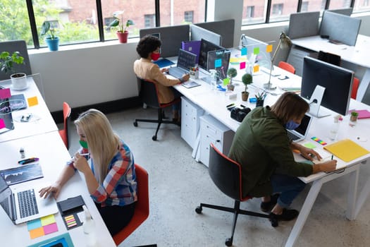 Diverse group of business people wearing face masks in creative office. group of people sitting at desks and using laptops. social distancing protection hygiene in workplace during covid 19 pandemic.