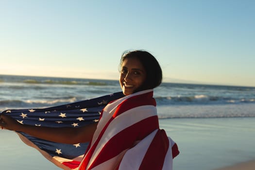 Portrait of mixed race patriotic woman with american flag on the beach looking to camera. healthy outdoor leisure time by the sea.