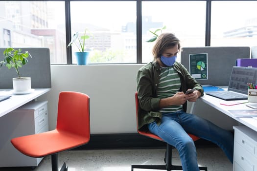 Caucasian businessman wearing face mask in creative office. woman sitting at desk, using smartphone. social distancing protection hygiene in workplace during covid 19 pandemic.