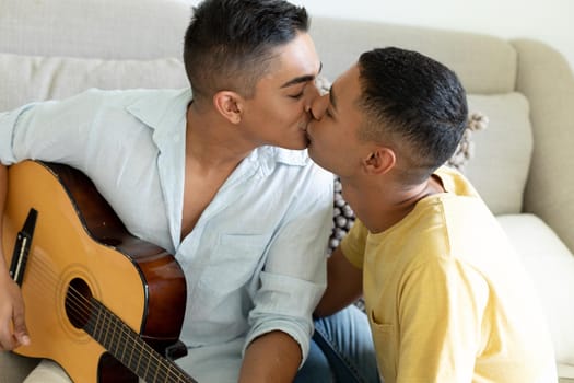 Diverse gay male couple sitting on sofa playing guitar and kissing. staying at home in isolation during quarantine lockdown.