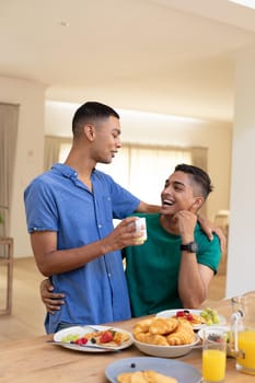 Diverse gay male couple sitting at table embracing. staying at home in isolation during quarantine lockdown.