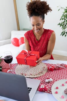 African american woman holding gift box on videocall at home. distant celebration of valentines day online and dating concept