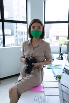Portrait of asian businesswoman wearing face mask holding camera in creative office. social distancing protection hygiene in workplace during covid 19 pandemic.