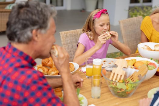 Caucasian girl and grandfather eating hamburgers at table with family having meal in the garden. three generation family sitting at table eating meal in the garden.