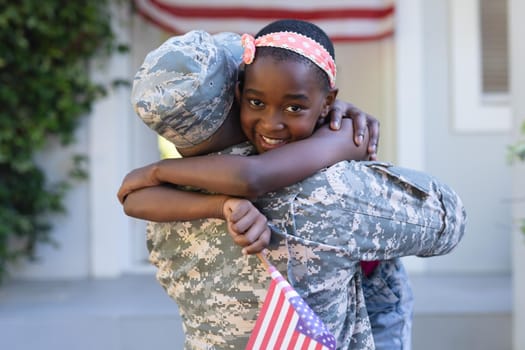 African american soldier father hugging smiling daughter holding flag in front of house. soldier returning home.