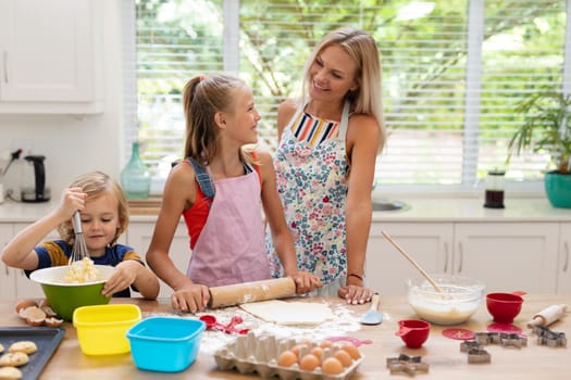Happy caucasian mother in kitchen with daughter and son, wearing aprons baking cookies together. staying at home in isolation during quarantine lockdown.