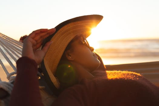 Mixed race woman on beach holiday lying in hammock relaxing during sunset. healthy outdoor leisure time by the sea.
