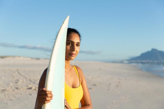 Happy mixed race woman carrying surfboard on the beach. healthy outdoor leisure time by the sea.