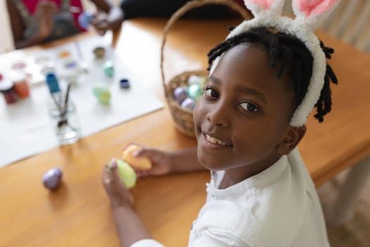 Happy african american boy wearing bunny ears painting colourful eggs. celebrating easter at home in isolation during quarantine lockdown.