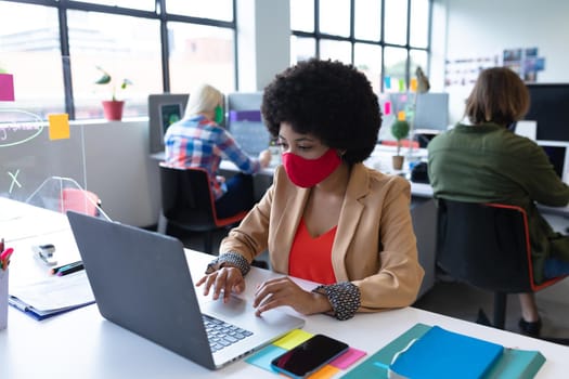 Mixed race businesswoman wearing face mask in creative office. woman sitting at desk, using laptop computer. social distancing protection hygiene in workplace during covid 19 pandemic.