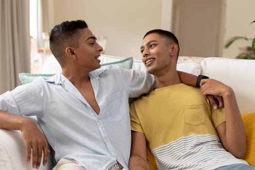 Diverse gay male couple sitting on sofa and embracing. staying at home in isolation during quarantine lockdown.