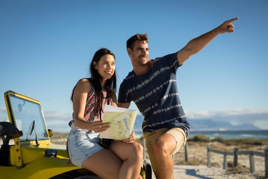 Happy caucasian couple sitting on beach buggy by the sea reading roadmap pointing. beach break on summer holiday road trip.