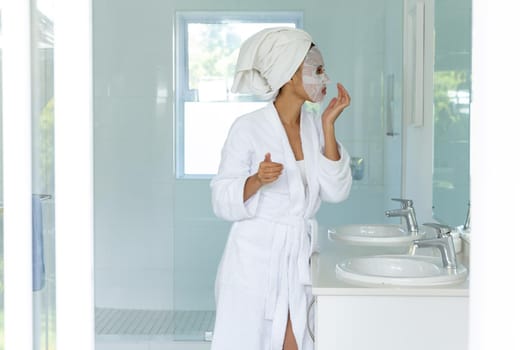 Mixed race woman wearing bathrobe and cleansing face mask in bathroom. staying at home in isolation during quarantine lockdown.