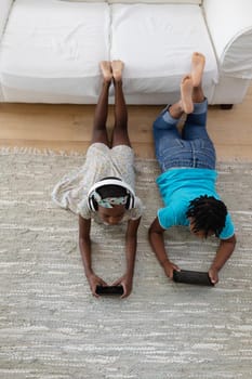 African american brother and sister wearing headphones lying on floor using smartphones. staying at home in isolation during quarantine lockdown.