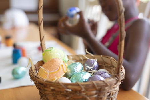 Basket of colourful eggs with african american girl painting in the background. celebrating easter at home in isolation during quarantine lockdown.