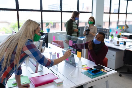 Two diverse business people wearing face masks in creative office. man and woman sitting at desk, discussing work. social distancing protection hygiene in workplace during covid 19 pandemic.