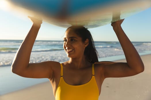 Happy mixed race woman on the beach carrying surfboard on head. healthy outdoor leisure time by the sea.