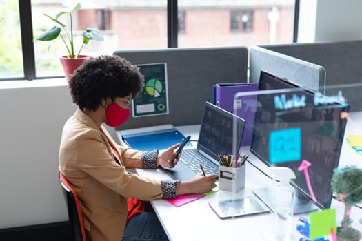 Mixed race businesswoman wearing face mask in creative office. woman sitting at desk, using laptop computer. social distancing protection hygiene in workplace during covid 19 pandemic.