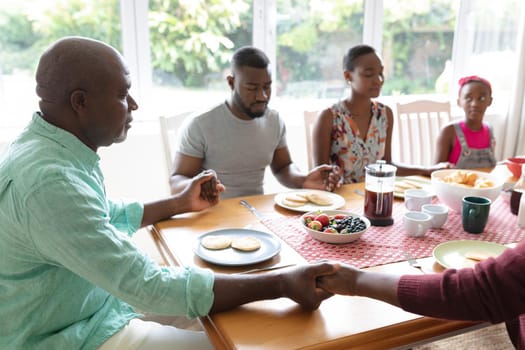 African american parents, daughter and grandparents sitting holding hands in prayer at dinner table. three generation family spending quality time together.