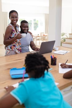Happy african american parents using laptop embracing holding coffee with kids working at the table. staying at home in isolation during quarantine lockdown.