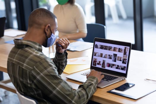 Diverse group of business people working in creative office. man wearing face mask and using laptop. social distancing protection hygiene in workplace during covid 19 pandemic.