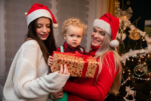 Parent with son in arms and sister give gift on Christmas Eve. Mother and daughter wearing a red Santa hat.