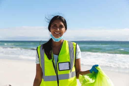 Portrait of smiling mixed race woman wearing face mask collecting rubbish from the beach. eco beach conservation during coronavirus covid 19 pandemic.
